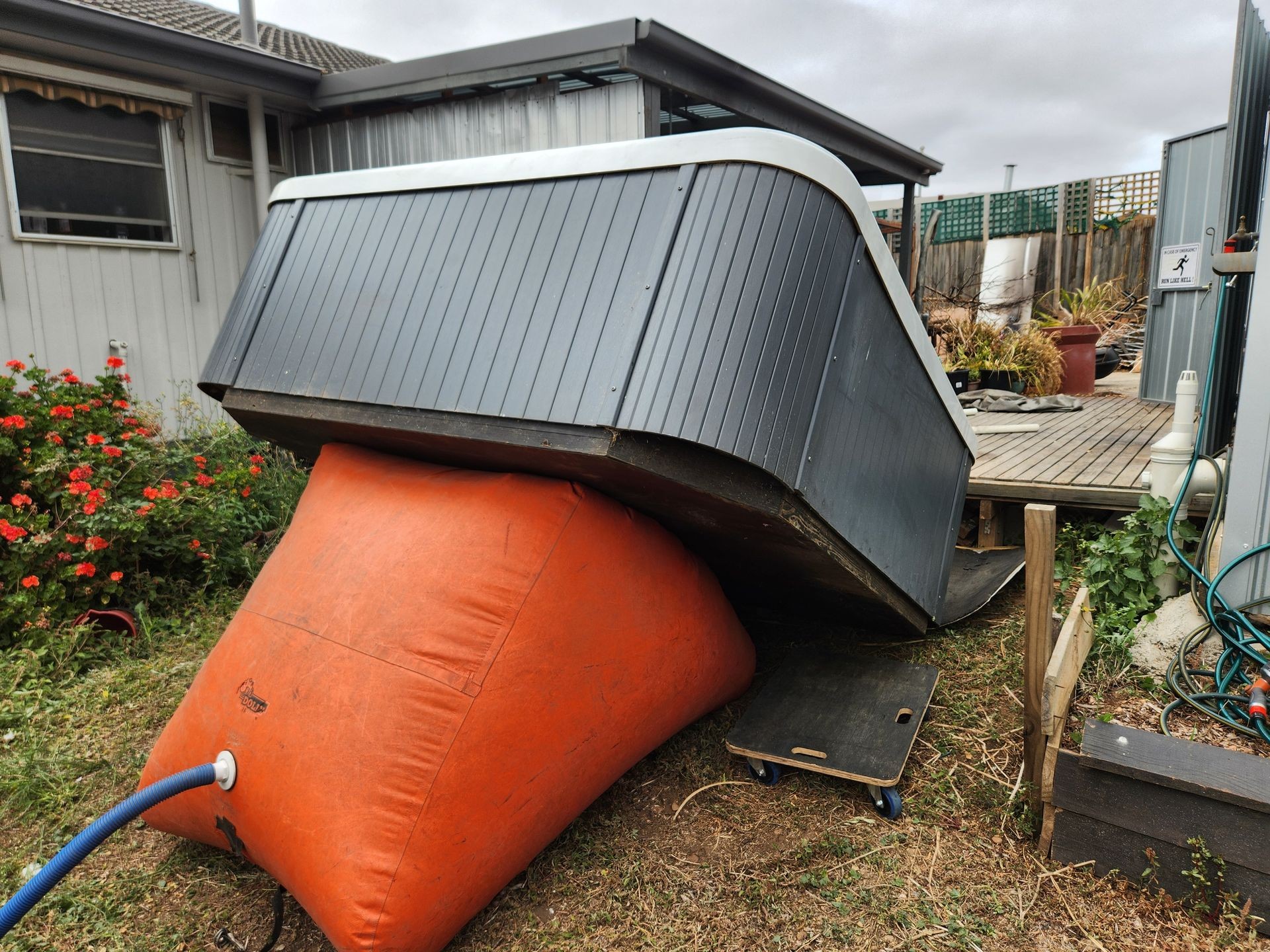 Tipped hot tub on orange airbag near backyard deck with plants and garden hose.